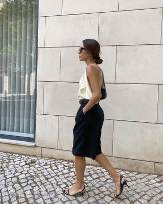 female fashion influencer Debora Rosa wears a low bun, silk camisole top, black Bermuda shorts, and simple black slingback sandals