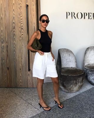 fashion influencer Tania Sarin poses in a black racerback tank top, Bottega Veneta black cloud clutch, white Bermuda shorts, and black flip-flop heeled sandals